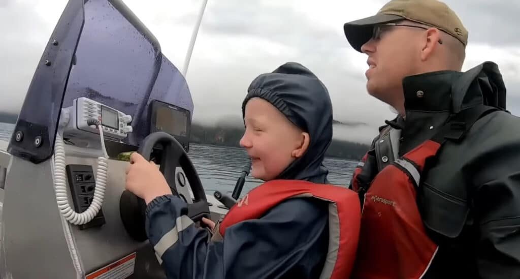 Luke Nichols of Outdoor Boys YouTuber teaches his son to steer a boat on a misty lake, both rocking life jackets and rain gear! This heartwarming moment captures their all-American bond before Luke’s YouTube exit. JV Political honors this MAGA dad’s legacy—share to celebrate family-first values!